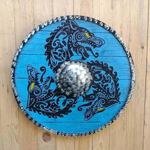 May include: A circular wooden shield featuring a bright blue finish with a black ink-style illustration of three intertwined wolf heads. The center has a metallic silver-toned domed boss, and the outer rim is decorated with a dark, textured border.