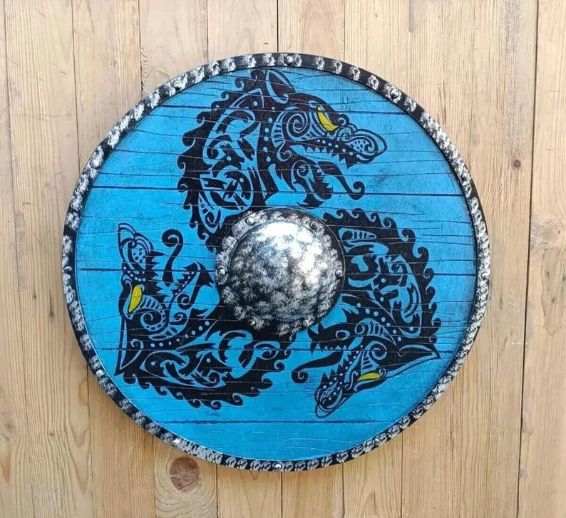 May include: A circular wooden shield featuring a bright blue finish with a black ink-style illustration of three intertwined wolf heads. The center has a metallic silver-toned domed boss, and the outer rim is decorated with a dark, textured border.
