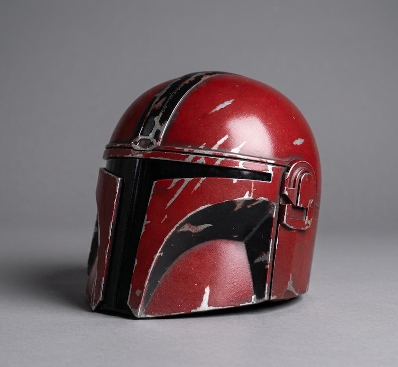 Puede incluir: Un casco rojo y negro con detalles plateados. El casco tiene un acabado brillante y un aspecto desgastado, con ara&ntilde;azos y marcas de desgaste. El dise&ntilde;o incluye una visera negra y una franja central. El casco es probablemente un accesorio o art&iacute;culo de colecci&oacute;n.
