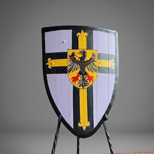 Op de afbeelding: Een schild in middeleeuwse stijl met een zwarte rand en een lichtpaarse achtergrond. Het schild heeft een gele kruis met een zwart-rood adelaarsymbleem in het midden. Zwarte verticale strepen en gouden accenten maken het ontwerp af.