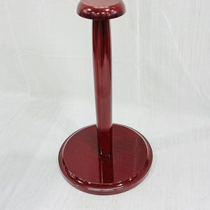 Puede incluir: Un expositor de sombreros de madera de color burdeos. Tiene una base circular, un poste delgado y una parte superior redondeada. La veta de la madera es visible y el acabado es brillante. Dise&ntilde;ado para sombreros.