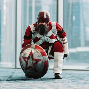 May include: A person in a red and white superhero costume, kneeling with a shield. The costume includes a helmet, chest emblem, and white boots. The shield is red, silver, and features a star.