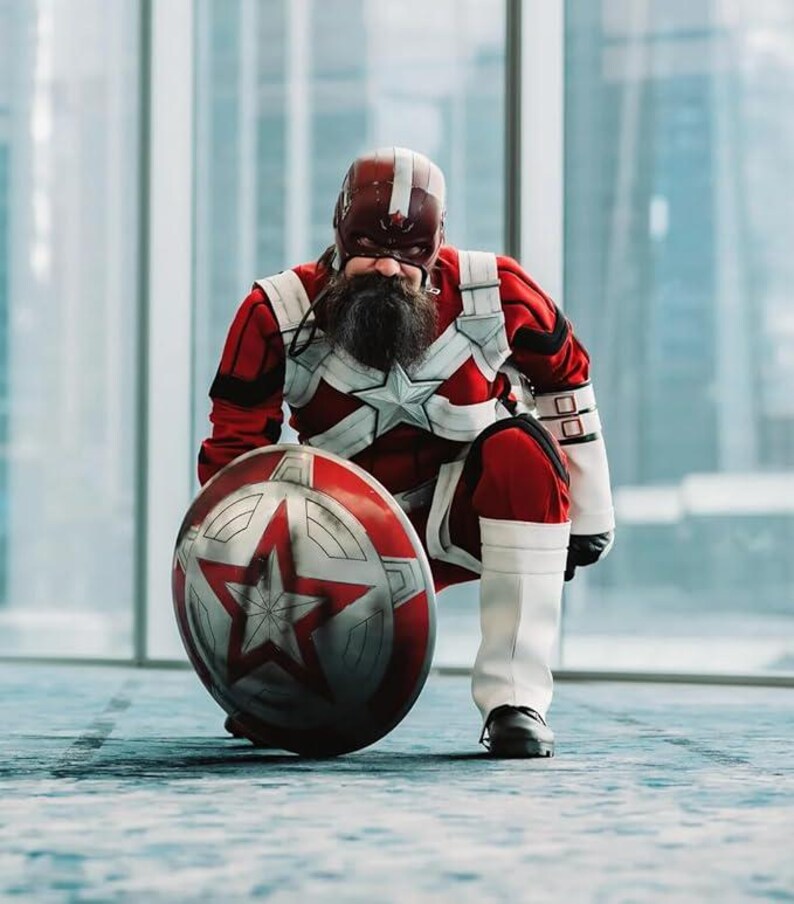 May include: A person in a red and white superhero costume, kneeling with a shield. The costume includes a helmet, chest emblem, and white boots. The shield is red, silver, and features a star.