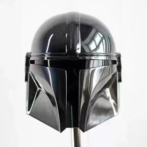 May include: A glossy black Mandalorian helmet with a sleek, futuristic design. The helmet features a rounded top with vertical ridges and a distinctive visor. The faceplate is angular and reflective, with a dark, shadowed appearance. The helmet is a prop or collectible.