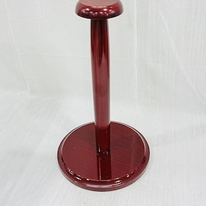 May include: A burgundy-colored wooden hat stand. The stand has a circular base, a tall, slender pole, and a rounded top. The wood grain is visible, and the finish is glossy. The stand is designed to hold hats.