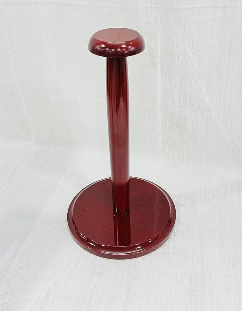 May include: A burgundy-colored wooden hat stand. The stand has a circular base, a tall, slender pole, and a rounded top. The wood grain is visible, and the finish is glossy. The stand is designed to hold hats.