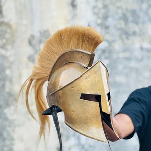 May include: A gold-colored Spartan helmet with a tan-colored plume. The helmet has a classic design with eye slits and a cheek guard. The plume is long and flowing, adding to the helmet's historical aesthetic. The helmet is held up against a neutral background.