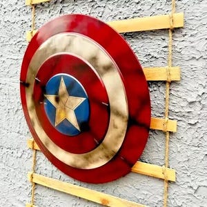 Op de afbeelding: Een rood, wit en goud schild met een blauw sterembleem. Het schild is gemonteerd op een houten ladder met touwsteunen, wat een decoratieve presentatie cre&euml;ert. Het ontwerp doet denken aan een superheld.