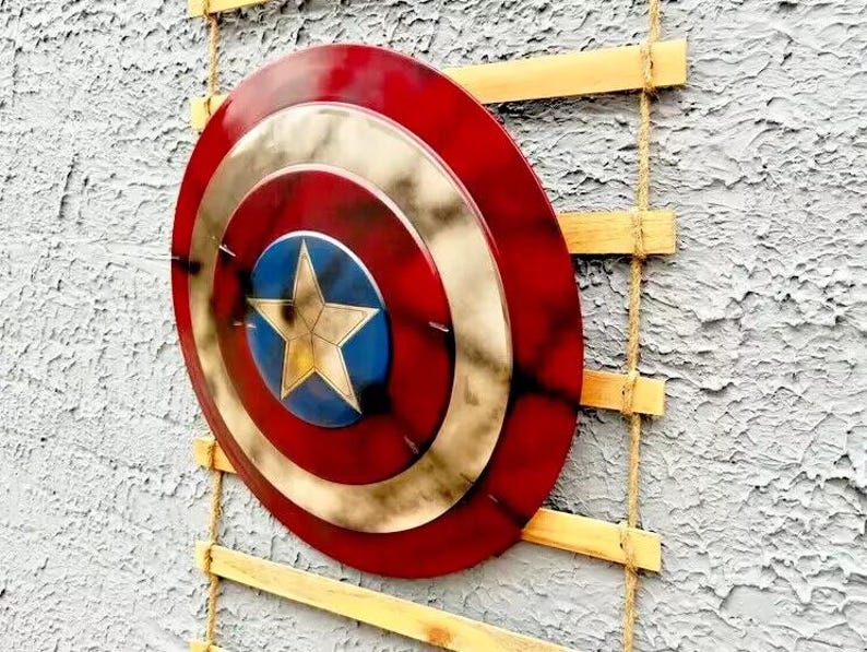 Op de afbeelding: Een rood, wit en goud schild met een blauw sterembleem. Het schild is gemonteerd op een houten ladder met touwsteunen, wat een decoratieve presentatie cre&euml;ert. Het ontwerp doet denken aan een superheld.