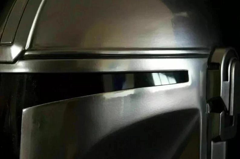 May include: Close-up of a metallic silver helmet with a visor. The helmet has a smooth, reflective surface with visible details and contours. The visor is dark, and the overall design suggests a futuristic or sci-fi theme.