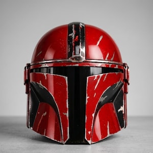 Puede incluir: Un casco mandaloriano rojo y negro con un acabado desgastado. El casco presenta una c&uacute;pula roja brillante y un &aacute;rea de visera negra. Los detalles plateados y desgastados por la batalla se suman al aspecto realista de este art&iacute;culo de colecci&oacute;n.