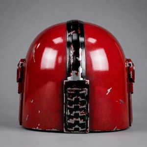 Puede incluir: Un casco rojo con una franja negra vertical en el centro y detalles negros. El casco tiene un aspecto desgastado con ara&ntilde;azos y desgaste visibles. El dise&ntilde;o incluye detalles laterales y una parte superior redondeada, sugiriendo un tema futurista o de ciencia ficci&oacute;n.