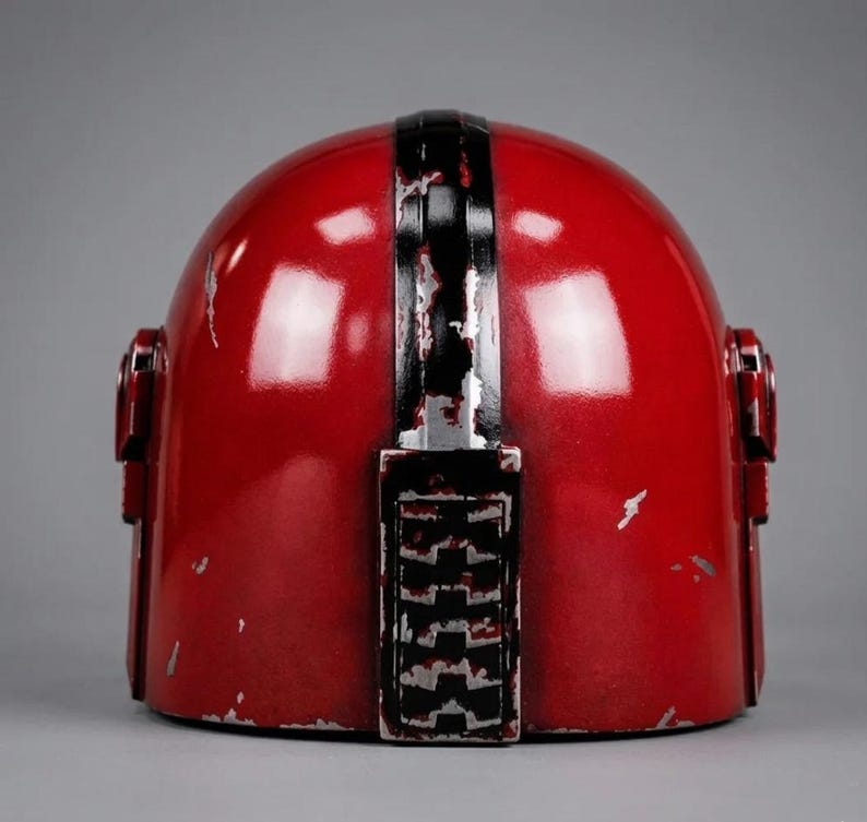 Puede incluir: Un casco rojo con una franja negra vertical en el centro y detalles negros. El casco tiene un aspecto desgastado con ara&ntilde;azos y desgaste visibles. El dise&ntilde;o incluye detalles laterales y una parte superior redondeada, sugiriendo un tema futurista o de ciencia ficci&oacute;n.