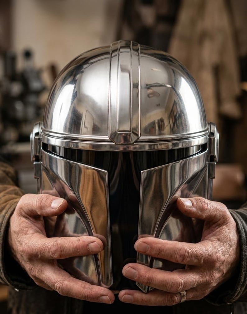 May include: A highly polished, silver-colored Mandalorian helmet. The helmet features a rounded top with vertical ridges and a distinctive visor design. The visor is dark, and the helmet is held by hands.