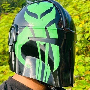May include: A black helmet with a glossy finish, featuring a vibrant green design. The helmet's design includes a stylized emblem on the front and sides. The helmet is a cosplay item.
