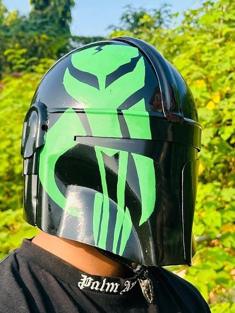 May include: A black helmet with a glossy finish, featuring a vibrant green design. The helmet's design includes a stylized emblem on the front and sides. The helmet is a cosplay item.