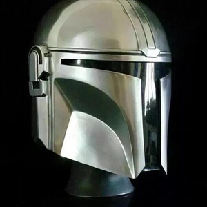 May include: A silver-colored Mandalorian helmet with a sleek, metallic finish. The helmet features a rounded top with a central ridge and a visor with a T-shaped opening. The design includes angular cheek plates and a dark visor area.