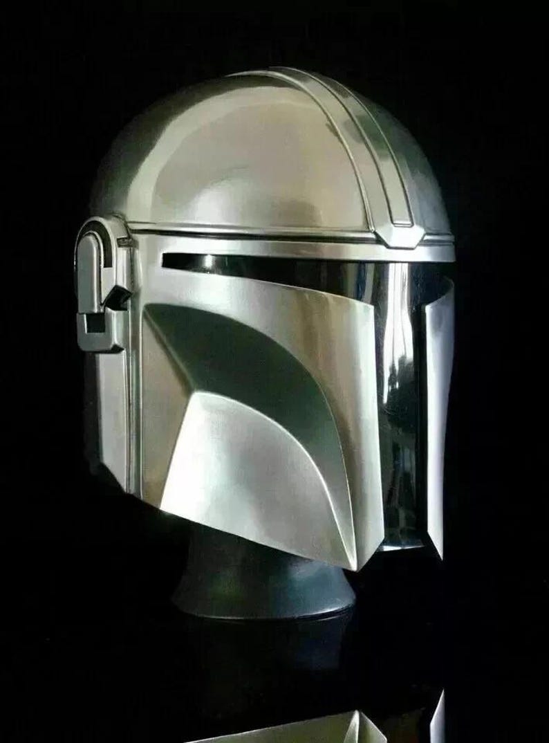 May include: A silver-colored Mandalorian helmet with a sleek, metallic finish. The helmet features a rounded top with a central ridge and a visor with a T-shaped opening. The design includes angular cheek plates and a dark visor area.