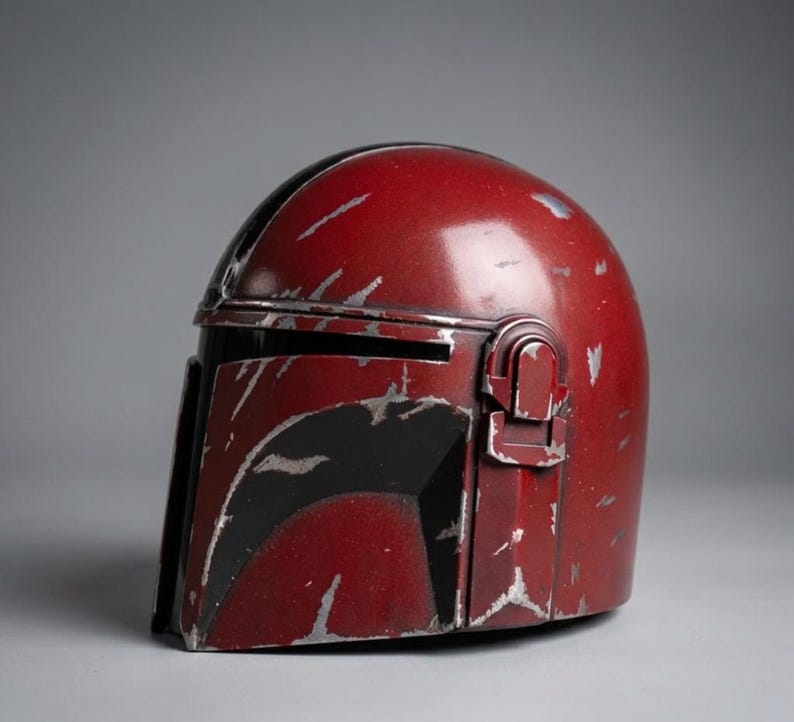 Puede incluir: Un casco rojo desgastado con ara&ntilde;azos plateados y detalles en negro. El casco tiene una visera y un accesorio lateral. El dise&ntilde;o sugiere un tema futurista o de ciencia ficci&oacute;n, posiblemente una pieza de disfraz o de colecci&oacute;n.