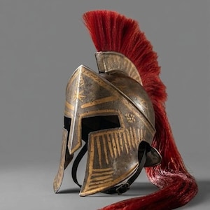 May include: A bronze-coloured Spartan helmet with gold detailing and a red plume. The helmet has a detailed design, a chin strap, and is set against a grey background. The helmet is a historical replica.