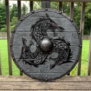 May include: A circular wooden shield featuring a grey weathered finish with horizontal plank detailing. The center displays a black Norse-style dragon motif with red eye accents and a prominent, hammered dark metal boss secured by rivets.