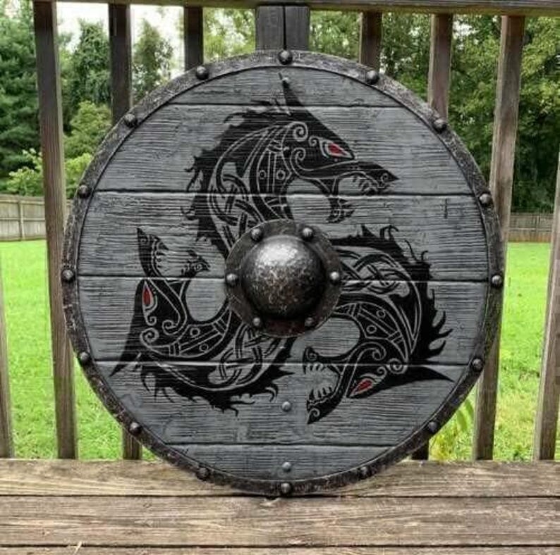 May include: A circular wooden shield featuring a grey weathered finish with horizontal plank detailing. The center displays a black Norse-style dragon motif with red eye accents and a prominent, hammered dark metal boss secured by rivets.