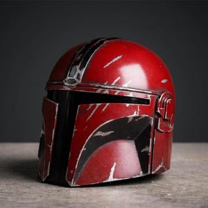 Puede incluir: Un casco mandaloriano rojo y negro con detalles plateados. El casco tiene un aspecto desgastado con ara&ntilde;azos y una visera negra. El casco es una r&eacute;plica del que usa el personaje de la serie Star Wars.