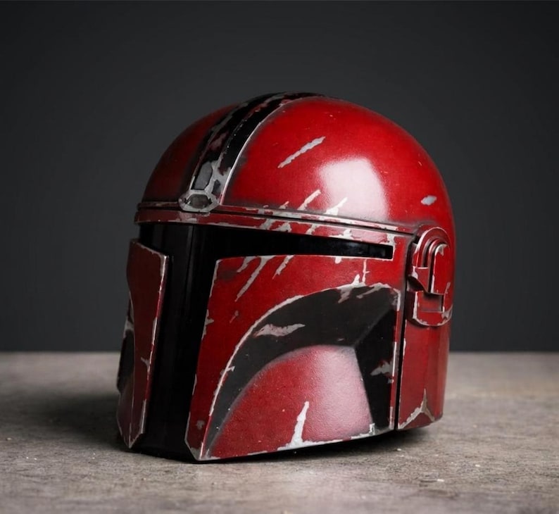 Puede incluir: Un casco mandaloriano rojo y negro con detalles plateados. El casco tiene un aspecto desgastado con ara&ntilde;azos y una visera negra. El casco es una r&eacute;plica del que usa el personaje de la serie Star Wars.