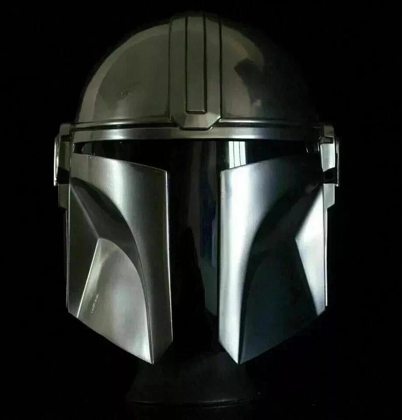 May include: A metallic silver Mandalorian helmet with a dark visor and a sleek, angular design. The helmet has a rounded top and a distinctive T-shaped visor, perfect for cosplay or display.