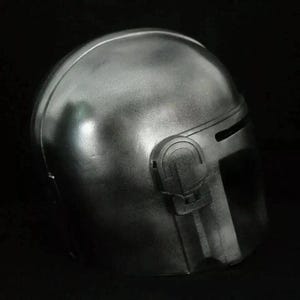 May include: A metallic silver helmet with a rounded top and a visor. The helmet has a dark, matte finish and features a prominent side detail with a rectangular shape. The overall design suggests a futuristic or sci-fi theme.
