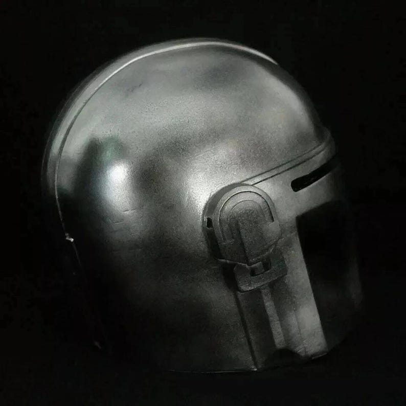 May include: A metallic silver helmet with a rounded top and a visor. The helmet has a dark, matte finish and features a prominent side detail with a rectangular shape. The overall design suggests a futuristic or sci-fi theme.
