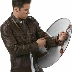 May include: A brown leather jacket with a zipper closure and multiple pockets. The person is holding a silver and red shield with brown leather straps. The shield has a circular design.