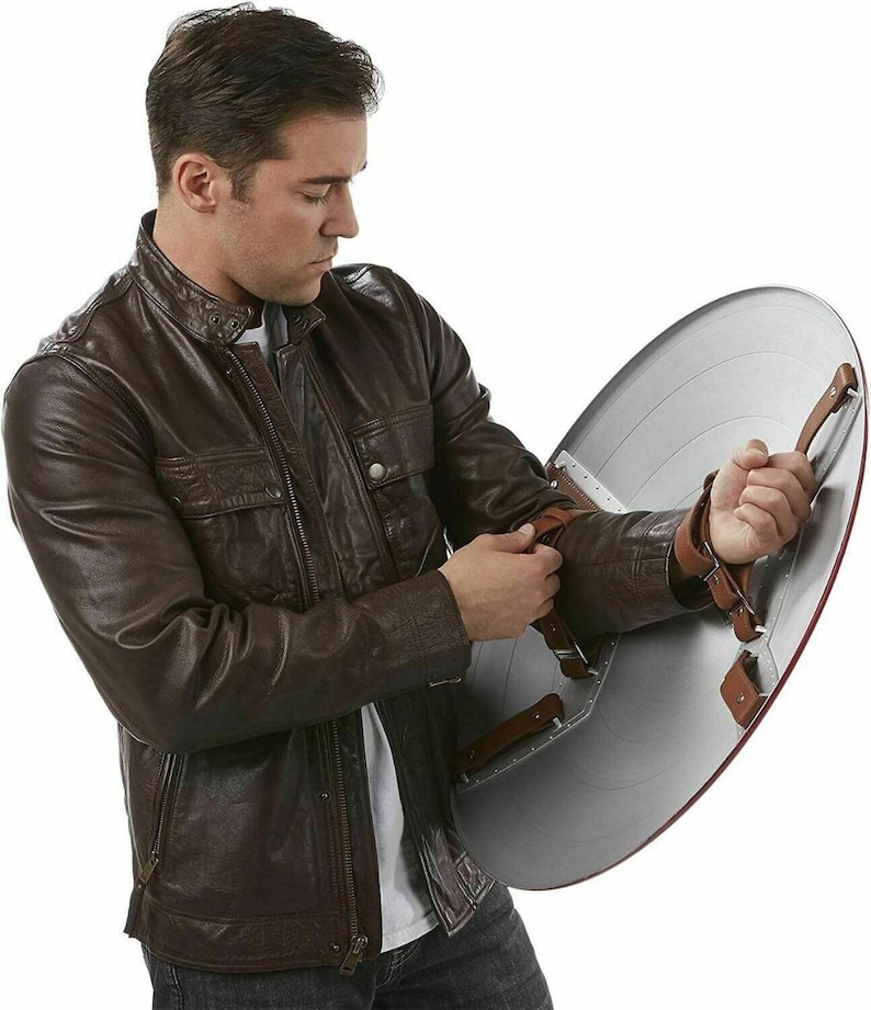 May include: A brown leather jacket with a zipper closure and multiple pockets. The person is holding a silver and red shield with brown leather straps. The shield has a circular design.