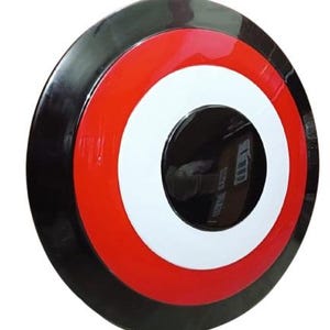May include: A round, glossy shield with a black outer rim, a red band, a white circle, and a black center. The shield is a prop or collectible item, likely inspired by a superhero or comic book.