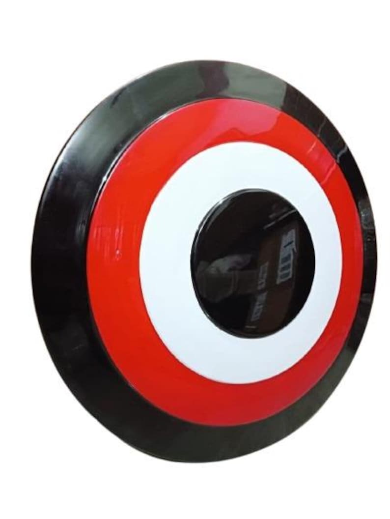 May include: A round, glossy shield with a black outer rim, a red band, a white circle, and a black center. The shield is a prop or collectible item, likely inspired by a superhero or comic book.