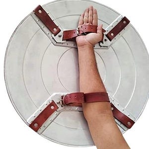Op de afbeelding: Een zilverkleurig, cirkelvormig schild met concentrische cirkels. Het heeft bruine leren riemen en metalen beugels voor bevestiging aan de arm. Het schild is ontworpen om op de arm te worden gedragen.