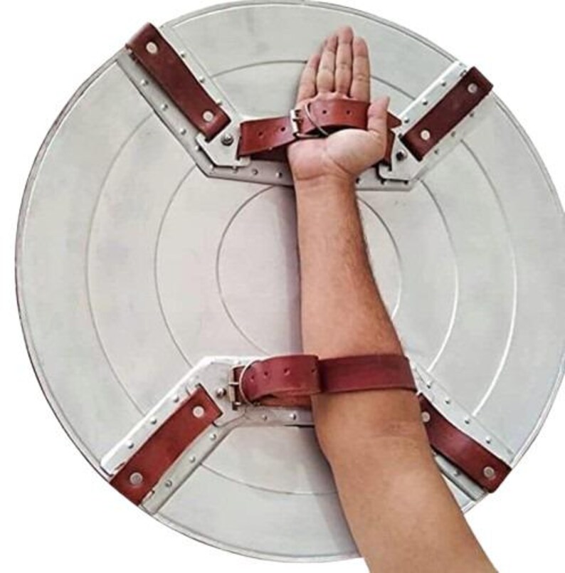 Op de afbeelding: Een zilverkleurig, cirkelvormig schild met concentrische cirkels. Het heeft bruine leren riemen en metalen beugels voor bevestiging aan de arm. Het schild is ontworpen om op de arm te worden gedragen.