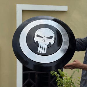 Op de afbeelding: Een cirkelvormig schild met een zwarte buitenring, een zilveren middelste ring en een zwarte binnencirkel. Het midden toont een opvallend zilveren schedel-embleem met langwerpige oogkassen en verticale tanden, het Punisher-logo.