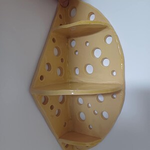 May include: A yellow, cheese-shaped corner shelf with three levels and numerous circular holes. The shelf is held up by a hand against a white wall. The shelf is made of ceramic or a similar material.