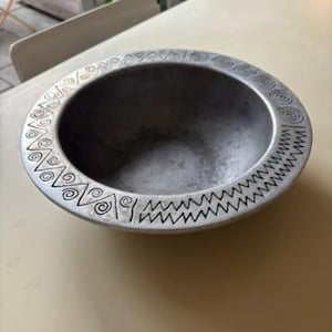 Könnte beinhalten: Eine silberfarbene, runde Schüssel mit einem geätzten Design am Rand. Der Rand weist ein sich wiederholendes Muster aus Wirbeln und Zickzacklinien auf. Die Schüssel steht auf einer hellen Oberfläche.