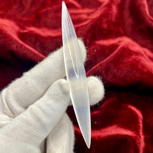 May include: A long, slender, translucent white selenite crystal wand with a smooth, polished surface and pointed ends. The crystal has a fibrous, reflective texture that catches the light, creating a subtle glow along its elongated, symmetrical shape.