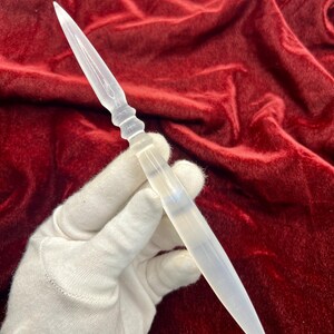 May include: A translucent white selenite crystal wand shaped like a double-pointed dagger. The object features a smooth, tapered blade on both ends with a central grip section that has subtle ridges, showcasing a natural, milky, and semi-opaque finish.