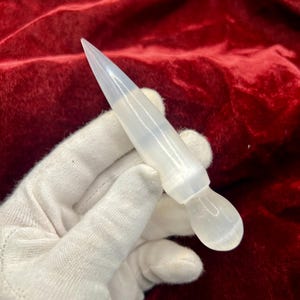 May include: A smooth, translucent white selenite crystal wand featuring a pointed, conical top and a rounded, bulbous base. The object has a polished, milky appearance with subtle internal striations visible throughout its elongated structure.