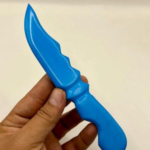 Può includere: Un coltello giocattolo di colore blu brillante, caratterizzato da una lama curva con dorso seghettato e un manico sagomato. L'oggetto presenta una finitura liscia e opaca e un design monoblocco continuo dalla punta fino alla base del manico.