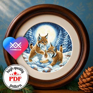 Op de afbeelding: Een ingelijst kruissteekpatroon met twee lynxwelpen die spelen in een besneeuwd bos onder een volle maan. Het kunstwerk is geplaatst in een ronde, donkere houten lijst. De afbeelding bevat een logo "high quality PDF pattern".