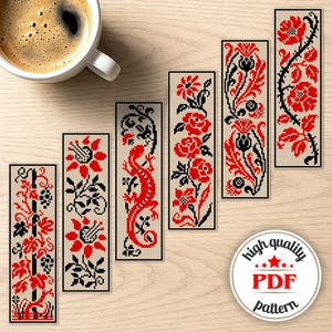 May include: A collection of six rectangular bookmarks with cross-stitch designs. Each bookmark features a different pattern in red and black on a beige background. The designs include floral motifs and a lizard. A cup of coffee sits in the upper left corner.