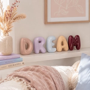Op de afbeelding: Een set van vijf individuele keramische letters die het woord DREAM vormen, elk met een gladde, glanzende, afgeronde ballonvorm in verschillende kleuren: oudroze, lavendel, lichtblauw, bleekgeel en donkerbordeaux.