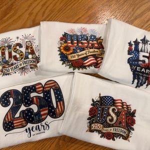 May include: Five white decorative towels with patriotic designs. Each towel features the text "USA" and "250 Years of Freedom" with American flag and firework graphics. The towels are folded and arranged on a wooden surface.
