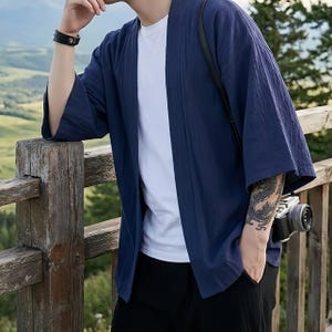 以下が含まれることがあります： ネイビーブルーの、袖が広い着物風ジャケット。オープンフロントのジャケットは、白いTシャツと黒いパンツの上に着用されています。黒いカメラがストラップから吊り下げられています。人物は木の柵に寄りかかっています。