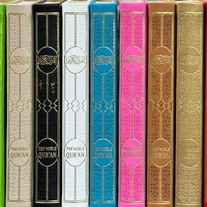 May include: A collection of vibrantly coloured books with ornate spines. The spines are in shades of blue, green, black, white, teal, pink, brown, gold, red, and grey. Each book features a circular emblem and the text "THE NOBLE QUR'AN".